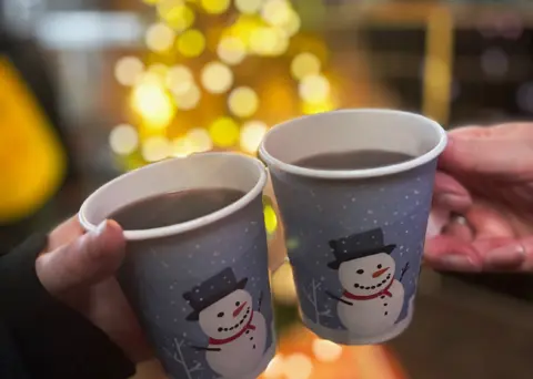 Zwei Personen halten einen Becher mit Glühwein vor einem Weihnachtsbaum.