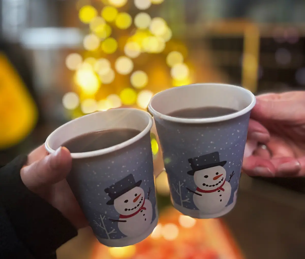 Zwei Personen halten einen Becher mit Glühwein vor einem Weihnachtsbaum.